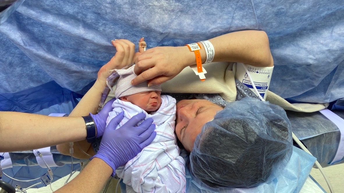 Newborn Luke swaddled in a hospital blanket, held close to Annie's face in the operating room